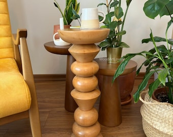 May include: A wooden side table with a unique design featuring a series of stacked rounded and triangular shapes. The table is made of natural wood and has a light brown finish. A white ceramic mug sits on the table's top surface.