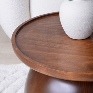 Puede incluir: Una mesa auxiliar redonda de madera marr&oacute;n oscuro con una superficie lisa y pulida. La mesa tiene un borde ligeramente elevado y una base redondeada. Un jarr&oacute;n blanco texturizado est&aacute; sobre la mesa. Un mueble elegante.