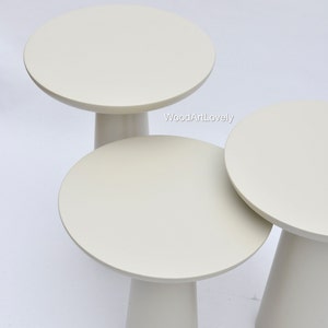 Op de afbeelding: Drie witte nesttafels met ronde bladen en taps toelopende poten. De tafels zijn op elkaar gestapeld, met de grootste tafel onderaan en de kleinste tafel bovenaan. WoodArtLovely is gedrukt op de grootste tafel.