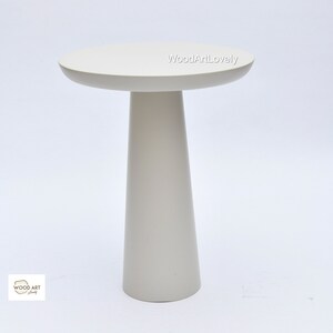 Op de afbeelding: Een witte bijzettafel met een rond blad en een hoge, taps toelopende poot. De tafel is gemaakt van hout en heeft een gladde, gepolijste afwerking.
