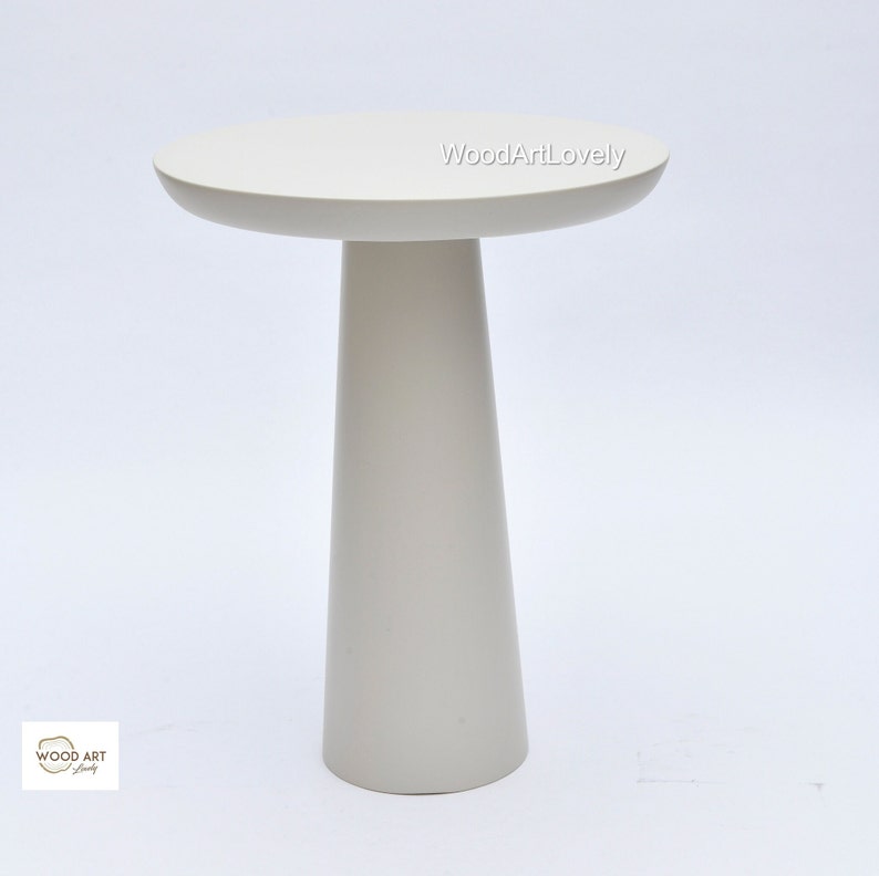 Op de afbeelding: Een witte bijzettafel met een rond blad en een hoge, taps toelopende poot. De tafel is gemaakt van hout en heeft een gladde, gepolijste afwerking.