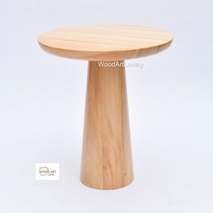 Op de afbeelding: Een ronde houten bijzettafel met een breed, vlak blad en een enkel, dik, taps toelopend been. De tafel is gemaakt van licht hout en heeft een natuurlijke afwerking.
