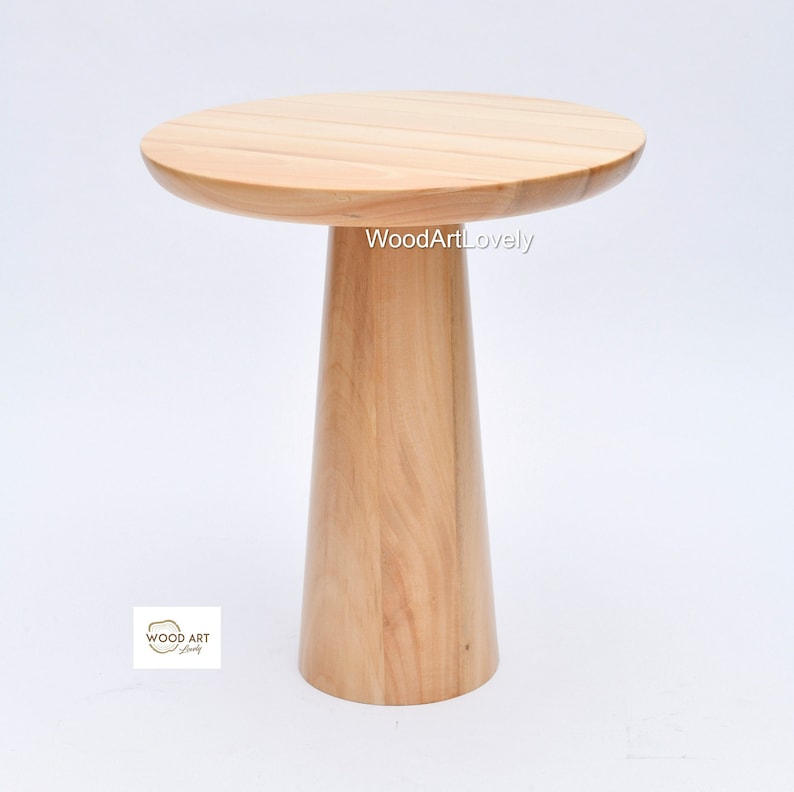 Op de afbeelding: Een ronde houten bijzettafel met een breed, vlak blad en een enkel, dik, taps toelopend been. De tafel is gemaakt van licht hout en heeft een natuurlijke afwerking.