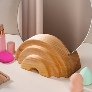 May include: A wooden stand shaped like a rainbow with a round mirror on top. The stand is made of light brown wood and has a natural finish. The mirror is clear and reflects the surrounding area.