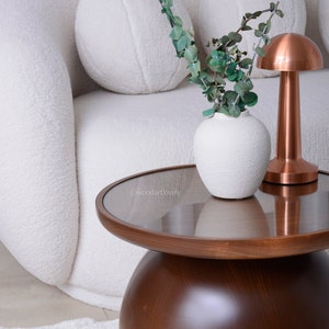Puede incluir: Escena de sal&oacute;n moderno con una mesa de centro redonda de madera oscura con tapa de cristal. Un jarr&oacute;n blanco texturizado contiene ramas de eucalipto verdes. Una l&aacute;mpara de cobre en forma de hongo est&aacute; sobre la mesa, junto a un sof&aacute; blanco curvado.