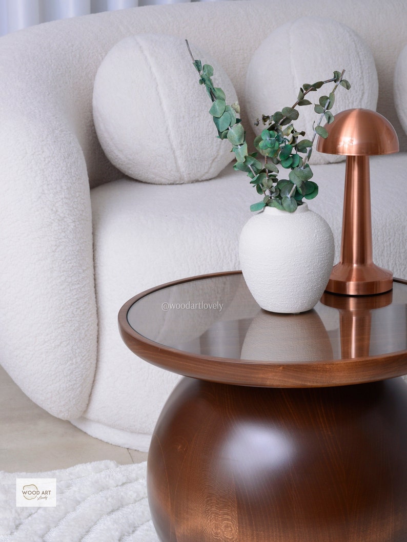 Puede incluir: Escena de sal&oacute;n moderno con una mesa de centro redonda de madera oscura con tapa de cristal. Un jarr&oacute;n blanco texturizado contiene ramas de eucalipto verdes. Una l&aacute;mpara de cobre en forma de hongo est&aacute; sobre la mesa, junto a un sof&aacute; blanco curvado.
