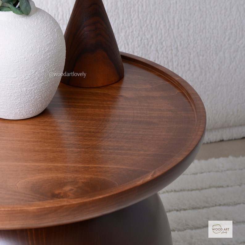 Puede incluir: Una mesa de centro redonda de madera con un acabado marr&oacute;n intenso. La mesa tiene un borde elevado y una base de pedestal. Un jarr&oacute;n blanco texturizado y un cono de madera marr&oacute;n oscuro est&aacute;n sobre la mesa. La habitaci&oacute;n es luminosa y tiene una alfombra blanca.