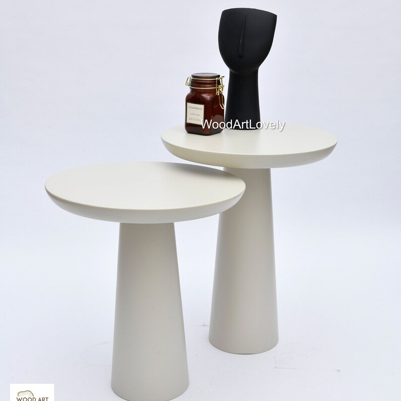 Op de afbeelding: Twee witte nesttafels met ronde bladen en taps toelopende poten. De tafels zijn op elkaar gestapeld, met een klein potje op de bovenste tafel. De tekst "WoodArtLovely" is zichtbaar op de bovenste tafel.