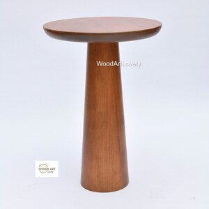 Op de afbeelding: Een houten bijzettafel met een rond blad en een hoog, taps toelopend poot. De tafel is gemaakt van lichtbruin hout en heeft een gladde afwerking. De tekst "WoodArtLovely" is zichtbaar op de tafel.