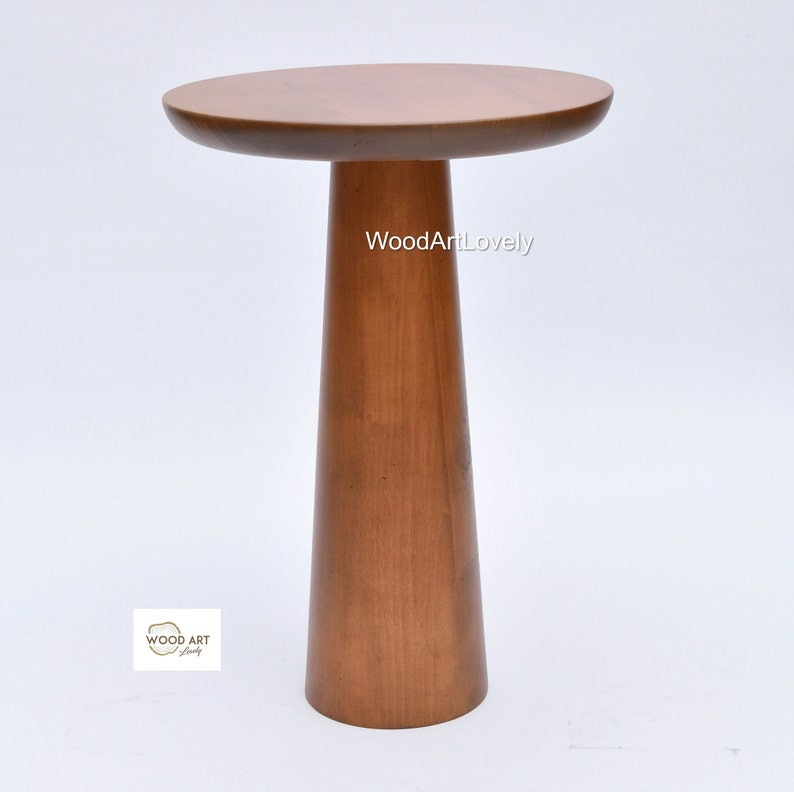 Op de afbeelding: Een houten bijzettafel met een rond blad en een hoog, taps toelopend poot. De tafel is gemaakt van lichtbruin hout en heeft een gladde afwerking. De tekst "WoodArtLovely" is zichtbaar op de tafel.