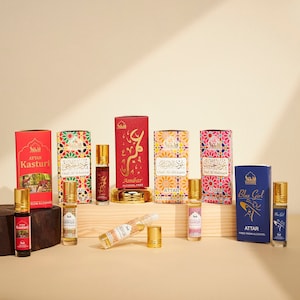 May include: A collection of ten small glass bottles of perfume oil with roll-on applicators. The bottles are arranged on a wooden surface. The bottles are decorated with colourful labels and gold accents. The labels feature Arabic script and the words "Attar" and "Alcohol-Free".