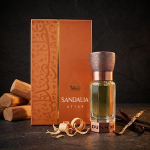May include: A rectangular orange box labeled SANDALIA ATTAR with Arabic calligraphy and a glass perfume bottle with a textured copper-colored cap and base. The bottle features the text DUKHNI at the bottom, accompanied by wood shavings.