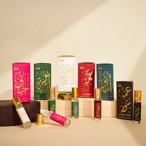 May include: An assortment of perfume bottles and boxes. The boxes are in various colors, including pink, green, red, and teal, with gold Arabic script. The perfume bottles are small, cylindrical, and filled with colored liquid. The text on the boxes includes "Attar Hayati" and "Ambar Oud."