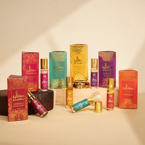 May include: An assortment of Sandalia attar perfume bottles and boxes. The bottles are small, cylindrical, and have gold caps. The boxes are in various colors, including red, gold, teal, and purple, with gold ornate designs and Arabic text.