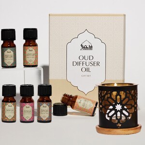 May include: A gift set of six small brown glass bottles with colorful labels containing oud diffuser oil. The bottles are arranged in two rows of three. The set also includes a black metal oil burner with a decorative design and a wooden base. The box is beige with gold accents and the text "DUKHNI OUD DIFFUSER OIL GIFT SET".