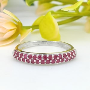 Peut inclure: Une bague en argent ornée de deux rangées de petites pierres précieuses rondes et rouges. La bague est posée sur un fond blanc, avec des feuilles et des fleurs vertes et jaunes floues en arrière-plan.