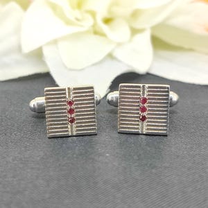 May include: A pair of silver cufflinks with a textured design and three red gemstones in each.