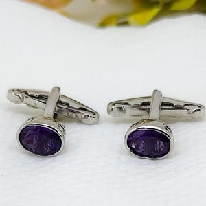 May include: A pair of silver cufflinks featuring oval, deep purple gemstones. The cufflinks are presented on a white surface, with blurred greenery in the background. The gemstones have a polished, reflective surface.