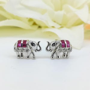 Può includere: Un paio di orecchini a lobo a forma di elefante in argento. Gli elefanti hanno accenti rettangolari color rubino sulla schiena e piccoli accenti a forma di diamante. Gli orecchini sono su uno sfondo sfocato.