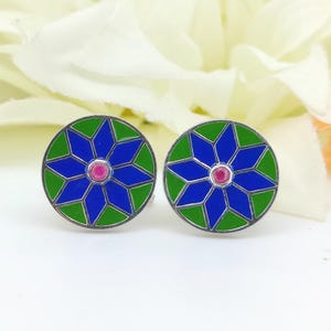 May include: A pair of round cufflinks with a floral design. The cufflinks feature a blue starburst pattern with green and silver accents, and a small pink center stone. The background is a soft white.