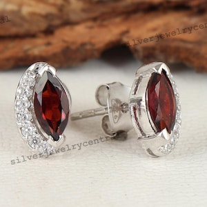 May include: A pair of silver stud earrings. Each earring features a marquise-cut, deep red gemstone surrounded by a halo of small, clear gemstones. The earrings have a post and butterfly backing.