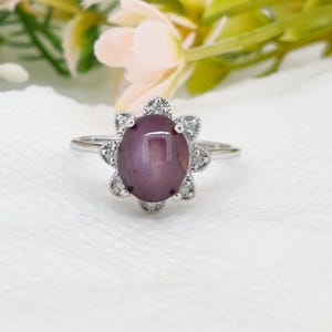 Op de afbeelding: Een zilveren ring met een grote, ovale, paarsrode edelsteen, omringd door kleine, heldere stenen in een bloemmotief. De ring heeft een dunne zilveren band en een gedetailleerde zetting.