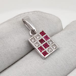 Op de afbeelding: Een zilveren hanger met een rechthoekig ontwerp. De hanger heeft een rasterpatroon van vierkante rode edelstenen en kleine, ronde, heldere edelstenen. De hanger wordt getoond tegen een lichtgrijze achtergrond.