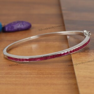 May include: A silver bangle bracelet with a row of red square-shaped gemstones. The bracelet is on a wooden surface.