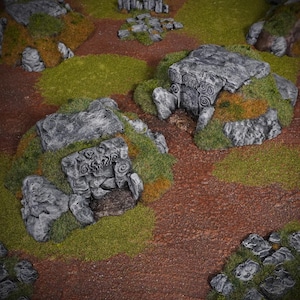 May include: A detailed tabletop terrain set featuring miniature stone structures with intricate carvings, surrounded by patches of green and brown vegetation. The scene is set on a reddish-brown surface, creating a realistic, miniature landscape.