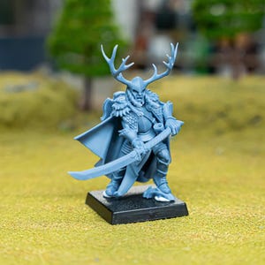 May include: A light blue miniature figurine of a warrior with antlers, a helmet, and a cape, holding a sword. The figurine is mounted on a black base and stands on a green surface. The background features blurred trees.