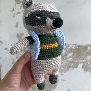 May include: A handmade crochet raccoon doll. The raccoon has a gray and white head with a black nose and eyes. It wears a green and yellow striped shirt and a light blue vest. The doll is held in a hand.