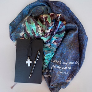 May include: A black journal with a silver cross necklace and a silver pen on top of it. The journal is lying on a blue and green patterned scarf with the text "what we hope for we do not see. Heb 11:1" printed on it.
