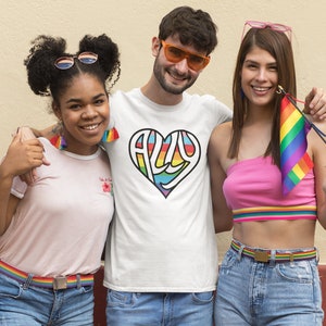 Puede incluir: Tres personas, dos mujeres y un hombre, están de pie juntos y sonriendo. El hombre lleva una camiseta blanca con un corazón arcoíris que dice "ALLY" en su interior. La mujer de la izquierda lleva una camiseta rosa y unos vaqueros azules con un cinturón arcoíris. La mujer de la derecha lleva un top rosa y unos vaqueros azules con un cinturón arcoíris. Ella sostiene una pequeña bandera arcoíris.