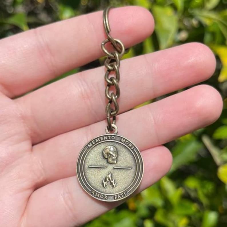 May include: Bronze-coloured keychain with a circular pendant. The pendant displays a skull over a flame, with the words "MEMENTO MORI" and "AMOR FATI". The keychain includes a chain and a keyring.