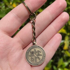 May include: Bronze-toned keychain with a circular pendant inscribed with "Wisdom", "Justice", "Temperance", and "Courage". The pendant is divided into quadrants with symbols. The keychain has a twisted chain and a ring.