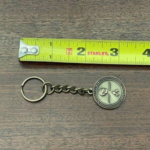 May include: A bronze-coloured keychain with a round pendant featuring a skull and bird design, with the words "MEMENTO MORI" and "AMOR FATI". The keychain is next to a yellow tape measure.