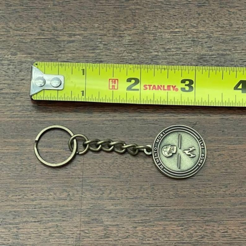 May include: A bronze-coloured keychain with a round pendant featuring a skull and bird design, with the words "MEMENTO MORI" and "AMOR FATI". The keychain is next to a yellow tape measure.