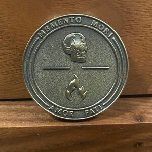 May include: A round, bronze-colored metal token with the words "Memento Mori" and "Amor Fati" inscribed around the edge. The token features a skull and a flame in the center.