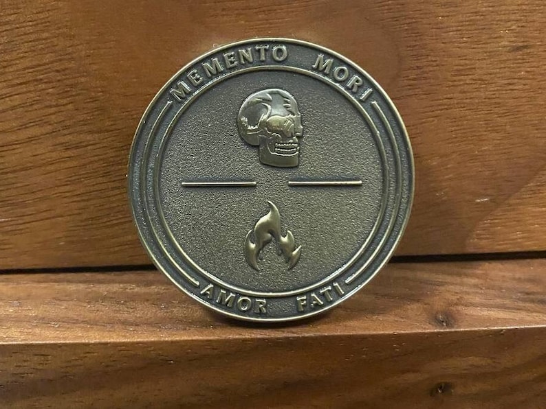 May include: A round, bronze-colored metal token with the words "Memento Mori" and "Amor Fati" inscribed around the edge. The token features a skull and a flame in the center.