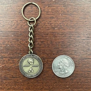 May include: Antique brass-coloured keychain with a round pendant featuring a skull, flame, and the words "MEMENTO MORI" and "AMOR FATI." The keychain includes a chain and a ring. A U.S. quarter is next to the keychain for size comparison.