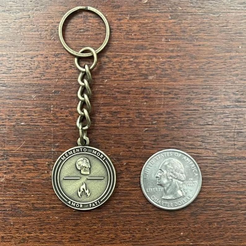 May include: Antique brass-coloured keychain with a round pendant featuring a skull, flame, and the words "MEMENTO MORI" and "AMOR FATI." The keychain includes a chain and a ring. A U.S. quarter is next to the keychain for size comparison.