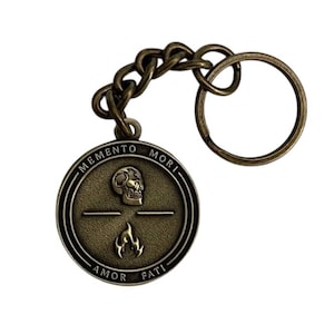 May include: Antique bronze keychain with a round pendant. The pendant features a skull, a flame, and the phrases "MEMENTO MORI" and "AMOR FATI". The keychain includes a chain and a key ring.