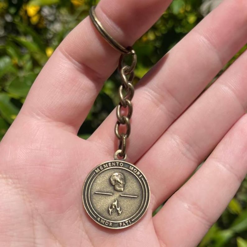 May include: Vintage brass keychain with a circular pendant. The pendant displays a skull, crosshairs, and flames, with the words "MEMENTO MORI" and "AMOR FATI" inscribed. The keychain includes a chain and a ring.