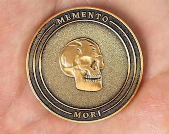 May include: A circular bronze-toned metal coin featuring a raised relief of a human skull in the center. The text MEMENTO is embossed along the top edge, and the text MORI is embossed along the bottom edge, both set against a dark background.