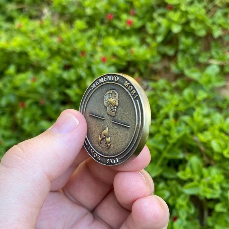May include: A gold-colored metal coin with a skull and flames design. The coin has the words "Memento Mori" and "Amor Fati" inscribed around the edge.