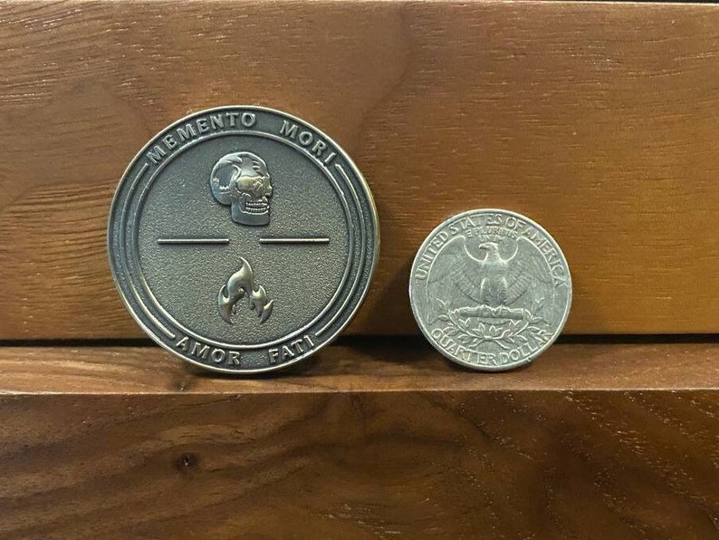 May include: A bronze coin with the words "Memento Mori" and "Amor Fati" inscribed around the edge. The coin features a skull and flames in the center.
