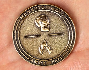 May include: A circular bronze-toned metal coin featuring an embossed skull at the top and a flame symbol at the bottom. The text MEMENTO MORI is inscribed along the upper edge, and AMOR FATI is inscribed along the lower edge of the coin.