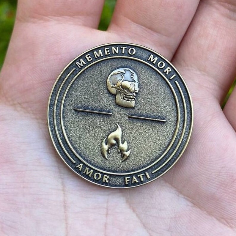 May include: A gold-toned round coin with the Latin phrase "Memento Mori" inscribed around the edge. The coin features a skull and a flame with the words "Amor Fati" inscribed below.