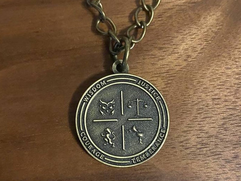 May include: A gold-toned pendant necklace with a round medallion featuring four symbols: an owl, a scale, a lion, and a cup. The medallion is inscribed with the words "Wisdom", "Justice", "Courage", and "Temperance".