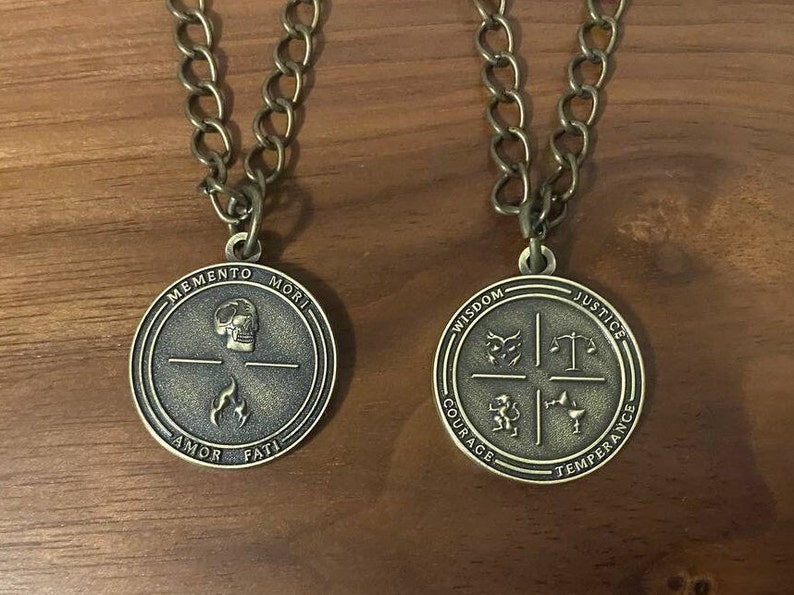 May include: Two gold-toned necklaces with round pendants. The pendant on the left has the words "Memento Mori" and "Amor Fati" inscribed around the edge. The pendant on the right has the words "Wisdom", "Justice", "Courage", and "Temperance" inscribed around the edge.